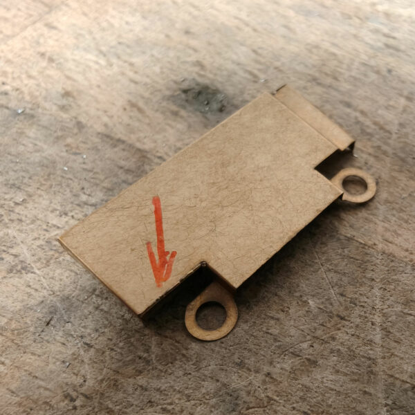 Petite pièce métallique plate avec une patte percée, présentant une soudure laser fine à l’angle. Une flèche rouge indique précisément la zone de soudure sur la surface posée sur un établi.